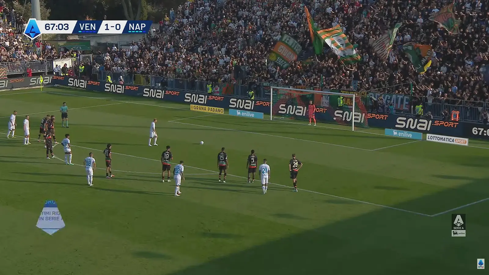 Il pallone sul dischetto del rigore durante Venezia-Napoli, con i giocatori fermi fuori area, il portiere sulla linea e i tifosi veneziani con le bandiere sugli spalti del Penzo